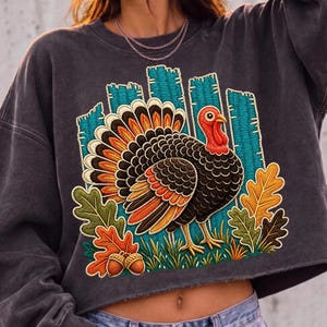 May include: A charcoal gray cropped sweatshirt featuring a large, colorful embroidered graphic of a turkey standing in grass. The design includes vibrant orange, yellow, and brown autumn leaves, two acorns, and a teal blue fence background.