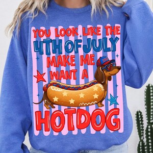 Op de afbeelding: Een blauwe sweater met ronde hals en een afbeelding van een teckel in een hotdogbroodje. De hond draagt een rood-wit-blauwe pet en een hartvormige zonnebril. De tekst luidt: YOU LOOK LIKE THE 4TH OF JULY MAKE ME WANT A HOTDOG, met sterren.