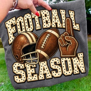 Puede incluir: Una camiseta gris oscuro con un estampado gr&aacute;fico que muestra el texto FOOTBALL SEASON en letras color crema con lunares negros. El dise&ntilde;o incluye un casco de f&uacute;tbol americano marr&oacute;n, un bal&oacute;n de f&uacute;tbol y un dedo de espuma marr&oacute;n hacia arriba.