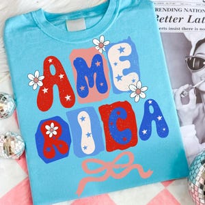 May include: A light blue t-shirt featuring the word AMERICA in large, colorful, blocky letters. The letters are filled with red, blue, and white patterns, including stars and small white flowers. A pink ribbon graphic is positioned below the text.