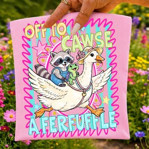 May include: Pink t-shirt with a whimsical illustration of a raccoon in overalls and a frog riding on a goose wearing a hat. The text on the shirt reads "OFF TO CAWSE A FERFUFFLE" in a playful font, surrounded by a decorative border.