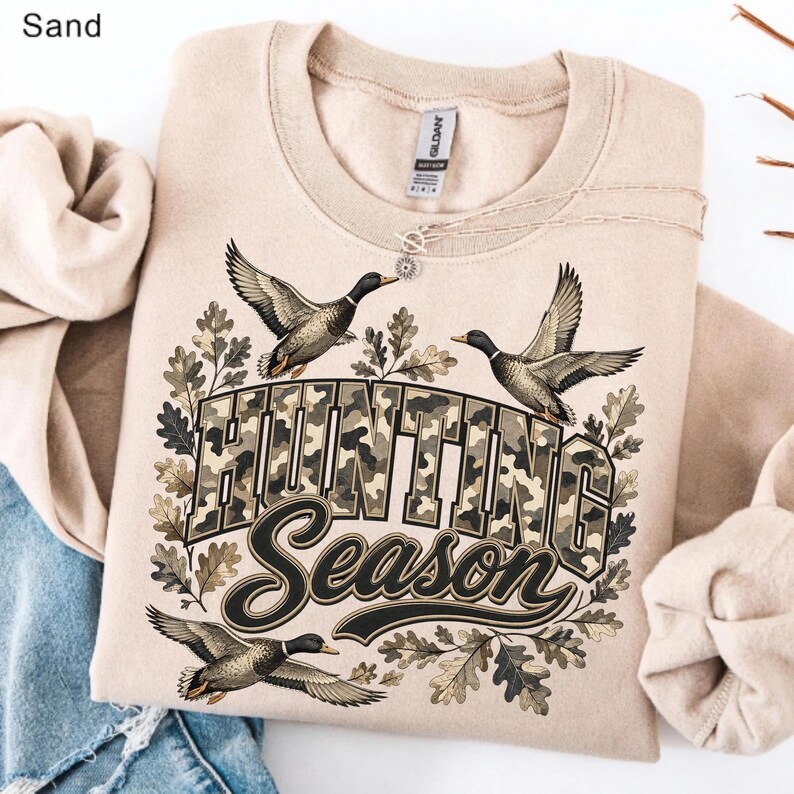 May include: A sand-colored crewneck sweatshirt featuring a graphic print of three flying ducks and oak leaves. The text "HUNTING Season" is displayed in the center with a camouflage pattern fill. The garment has a ribbed collar and long sleeves.
