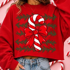 May include: A red long-sleeve sweatshirt featuring a central graphic of a candy cane with a large red bow. The background of the design consists of horizontal rows of green pine branch patterns. The sweatshirt has a crew neckline and a cropped hem.