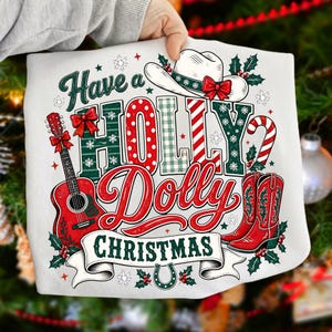 May include: A white sweatshirt featuring a festive graphic with the text Have a HOLLY Dolly CHRISTMAS. The design includes a cowboy hat, a red acoustic guitar, red cowboy boots, a candy cane, holly leaves, and a horseshoe on a banner.