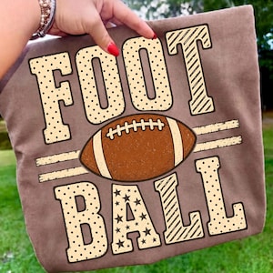 May include: A brown t-shirt featuring the text FOOTBALL in large, cream-colored block letters with polka dot and star patterns. A brown American football graphic with white laces is positioned horizontally between the words FOOT and BALL.