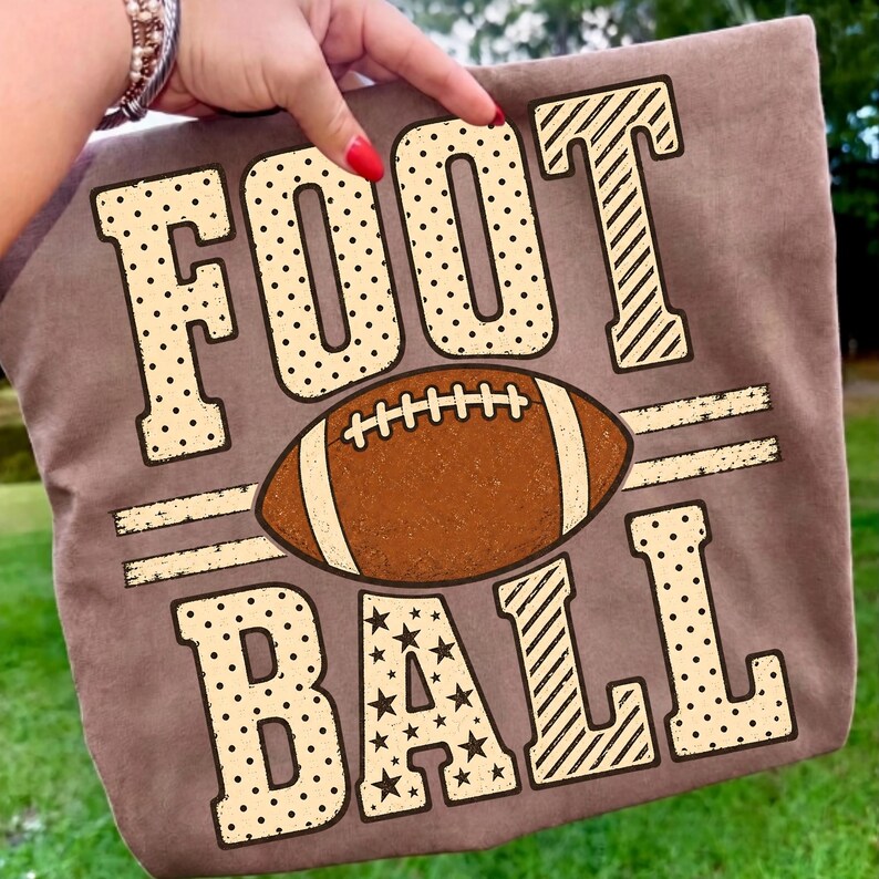 May include: A brown t-shirt featuring the text FOOTBALL in large, cream-colored block letters with polka dot and star patterns. A brown American football graphic with white laces is positioned horizontally between the words FOOT and BALL.