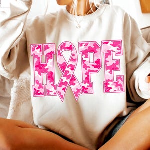 Peut inclure: Un sweat-shirt beige &agrave; col rond avec le mot HOPE imprim&eacute; en grandes lettres majuscules. La lettre O est remplac&eacute;e par un ruban de sensibilisation rose. Les lettres et le ruban pr&eacute;sentent un motif camouflage rose et blanc textur&eacute;.