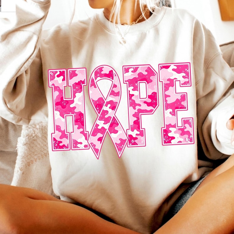 Peut inclure: Un sweat-shirt beige &agrave; col rond avec le mot HOPE imprim&eacute; en grandes lettres majuscules. La lettre O est remplac&eacute;e par un ruban de sensibilisation rose. Les lettres et le ruban pr&eacute;sentent un motif camouflage rose et blanc textur&eacute;.