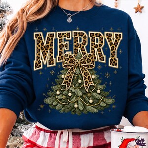 Op de afbeelding: Een marineblauw sweatshirt met lange mouwen en het woord MERRY in grote blokletters met een luipaardprint. Onder de tekst staat een afbeelding van een groene kerstboom versierd met gouden kralen, kleine witte ornamenten en een grote luipaardstrik.