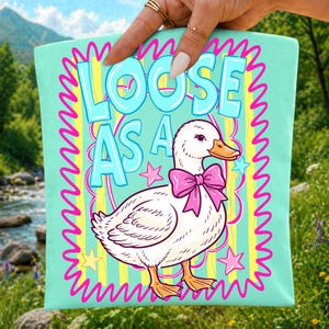 K&ouml;nnte beinhalten: Eine hellminzgr&uuml;ne Tragetasche mit einer wei&szlig;en Ente, die eine rosa Schleife um den Hals tr&auml;gt. Der Text LOOSE AS A GOOSE ist in gro&szlig;en blauen Buchstaben &uuml;ber der Ente gedruckt. Das Design enth&auml;lt gelbe vertikale Streifen, rosa Wellenr&auml;nder und Sterne.