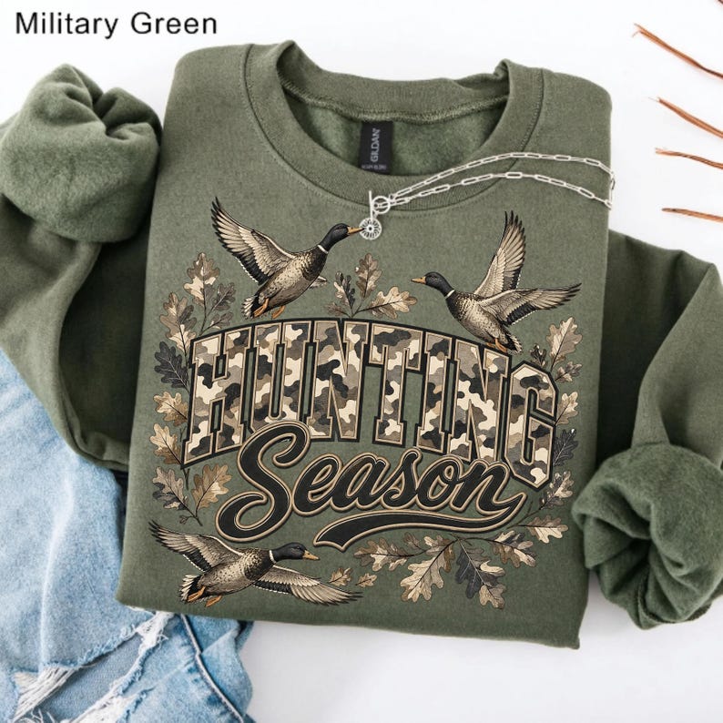 May include: A military green crewneck sweatshirt featuring a graphic print of three flying ducks and oak leaves. The text "HUNTING SEASON" is printed in a camouflage pattern with a dark outline. A silver chain necklace rests on the neckline.