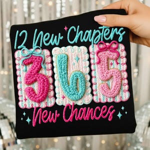 May include: Black t-shirt with the text "12 New Chapters 365 New Chances." The numbers 3, 6, and 5 are in a crochet style, with pink and teal colors. The text is in a cursive font, with a teal and pink color scheme.
