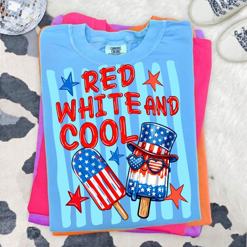 May include: A light blue t-shirt featuring the text RED WHITE AND COOL in red letters. The graphic includes a popsicle with a stars and stripes pattern and a second popsicle wearing a patriotic top hat and heart-shaped sunglasses with stars.