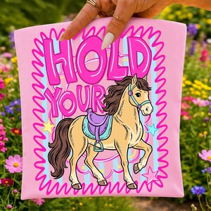 May include: A light pink fabric item featuring a graphic of a tan horse with a dark brown mane and tail, wearing a purple saddle. The text "HOLD YOUR" is printed in large, bold pink letters above the horse, surrounded by a wavy pink border.