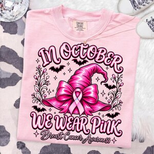 May include: A light pink t-shirt featuring a graphic of a pink witch hat adorned with a breast cancer awareness ribbon. The text reads In October We Wear Pink Breast Cancer Awareness, surrounded by black bats, stars, and decorative floral vines.