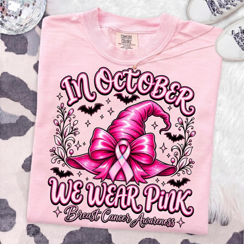 May include: A light pink t-shirt featuring a graphic of a pink witch hat adorned with a breast cancer awareness ribbon. The text reads In October We Wear Pink Breast Cancer Awareness, surrounded by black bats, stars, and decorative floral vines.