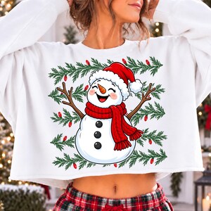 Puede incluir: Una sudadera blanca corta con el gr&aacute;fico de un mu&ntilde;eco de nieve sonriente que lleva una bufanda roja y un gorro de Pap&aacute; Noel rojo. El mu&ntilde;eco tiene brazos de rama y tres botones negros. El fondo del gr&aacute;fico incluye ramas de pino verdes y luces rojas.