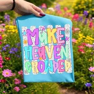 May include: A bright turquoise t-shirt featuring a graphic print with the text "MAKE HEAVEN CROWDED" in colorful, patterned bubble letters. The design includes black and white polka dot stripes, small stars, flowers, and pink ribbon accents.