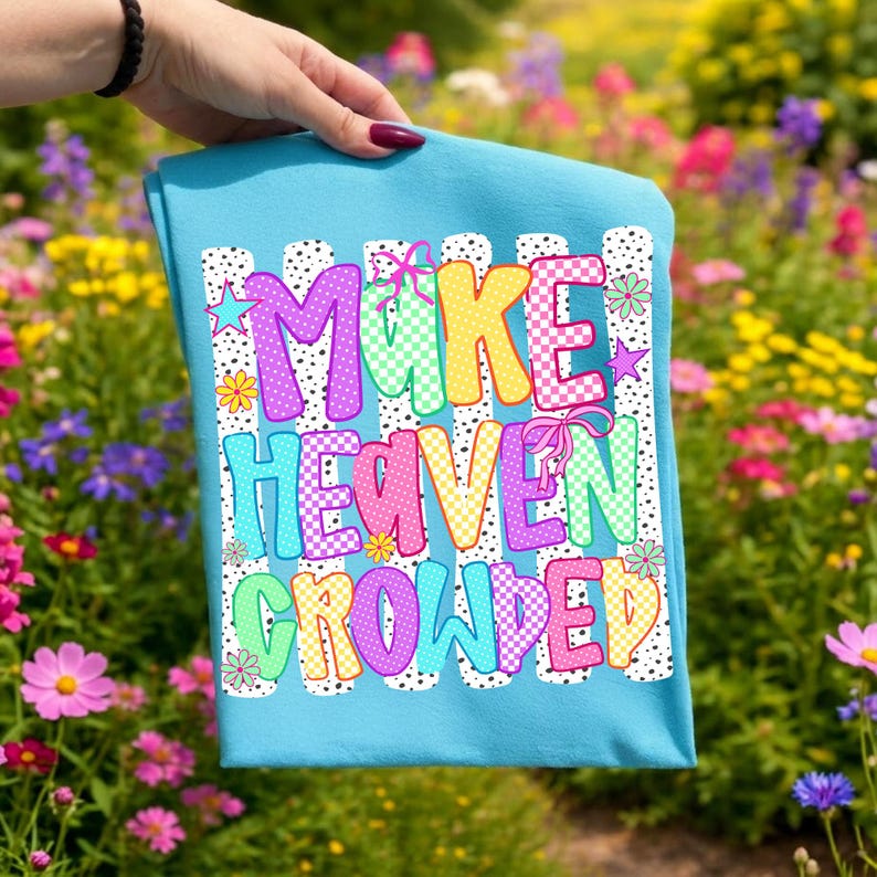 May include: A bright turquoise t-shirt featuring a graphic print with the text "MAKE HEAVEN CROWDED" in colorful, patterned bubble letters. The design includes black and white polka dot stripes, small stars, flowers, and pink ribbon accents.