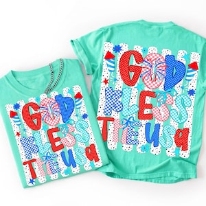 K&ouml;nnte beinhalten: Ein mintgr&uuml;nes T-Shirt mit einem Grafikdruck, der den Text "GOD BLESS THE USA" in rot-wei&szlig;-blauen Mustern zeigt. Das Design umfasst Feuerwerk, Sterne und Raketenmotive vor einem Hintergrund aus vertikalen schwarz-wei&szlig;en Punkten.