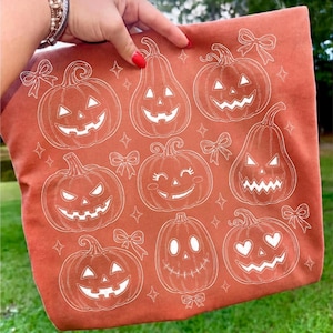 May include: A burnt orange fabric item featuring a repeating white line art pattern of various jack-o'-lanterns with different facial expressions, interspersed with small decorative bows and four-pointed star shapes across the surface.
