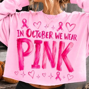 May include: A light pink long-sleeve sweatshirt featuring the text "IN OCTOBER WE WEAR PINK" in a bold, brushstroke-style font. The design includes several pink awareness ribbons, outlined hearts, and four-pointed star icons printed on the front.