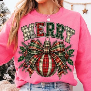 Puede incluir: Una sudadera rosa brillante de manga larga con la palabra MERRY en una fuente de estilo universitario con patr&oacute;n de cuadros. Debajo del texto hay un gr&aacute;fico de un adorno navide&ntilde;o con un lazo de cuadros, ramas de pino verdes y bayas rojas.