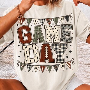 May include: A cream-colored t-shirt featuring the text GAME DAY in large block letters. The letters are decorated with football-themed patterns including field markings, stars, and plaid. Bunting flags with football icons border the text.