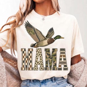 May include: A cream-colored t-shirt featuring a graphic of a flying mallard duck above the word MAMA. The letters are filled with distinct patterns: a brown and tan checkerboard, leopard print, green camouflage, and horizontal brown stripes.