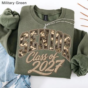 May include: A military green crewneck sweatshirt featuring the word SENIOR in a camouflage print with a black outline, and the text Class of 2027 in a tan script font below. A silver chain necklace rests on the neckline of the garment.