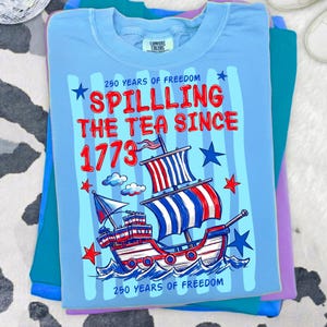 Puede incluir: Una camiseta azul claro con un gr&aacute;fico de un velero con velas a rayas rojas, blancas y azules. El texto dice 250 YEARS OF FREEDOM, SPILLING THE TEA SINCE 1773 y 250 YEARS OF FREEDOM, decorado con estrellas rojas y azules y nubes.