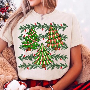 May include: A cream-coloured t-shirt featuring a graphic print of three stylised Christmas trees. The trees are decorated with red and white stripes, green and red plaid, and green and white stripes. Green pine branches and red lights surround them.
