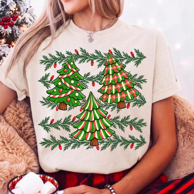 May include: A cream-coloured t-shirt featuring a graphic print of three stylised Christmas trees. The trees are decorated with red and white stripes, green and red plaid, and green and white stripes. Green pine branches and red lights surround them.