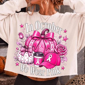 May include: A cream-colored long-sleeve sweatshirt featuring a graphic print with the text In October We Wear Pink. The design includes a large pink pumpkin with a bow, a smaller white pumpkin, a coffee cup, a baseball cap, and pink roses.