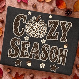 Może przedstawiać: Czarna prostokątna powierzchnia z napisem COZY SEASON wykonanym dużymi brązowymi literami w białe kropki. Centralnie umieszczona grafika dyni w panterkę znajduje się między słowami. Małe białe gwiazdki, kwiaty i żołędzie otaczają tekst.