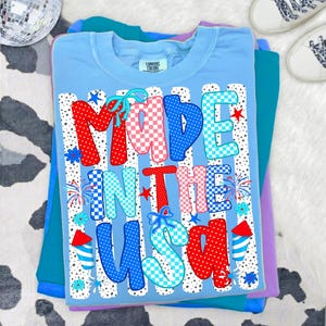 May include: A light blue t-shirt featuring the text MADE IN THE USA in large, multi-coloured, patterned letters. The design includes red, white, and blue polka dots, checkered patterns, and firework graphics, with small blue bows accenting the letters.