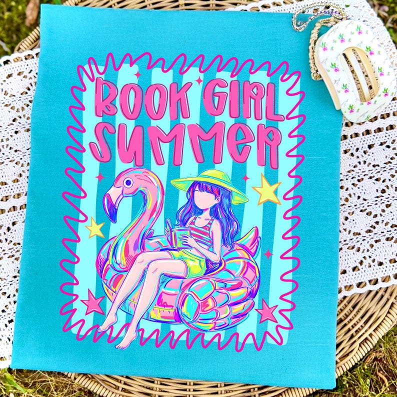 May include: A teal t-shirt featuring a graphic of a person in a yellow sun hat sitting on a pink flamingo pool float while reading a book. The text BOOK GIRL SUMMER is printed in pink above the illustration, surrounded by a wavy pink border.