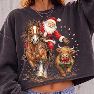 Puede incluir: Una sudadera corta de color gris carb&oacute;n con un estampado gr&aacute;fico de Santa Claus montando un caballo marr&oacute;n con una mancha blanca. A su lado, una vaca Highland peluda sostiene un regalo verde con un lazo rojo, rodeada de luces navide&ntilde;as.