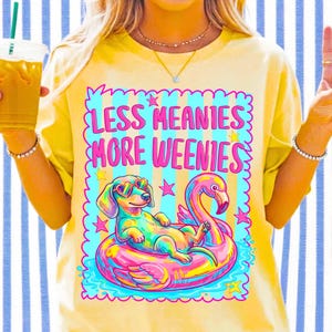 May include: A pale yellow t-shirt featuring a graphic of a dachshund wearing heart-shaped sunglasses while floating on a pink flamingo pool float. The text reads LESS MEANIES MORE WEENIES in pink letters, surrounded by a blue and pink scalloped border.