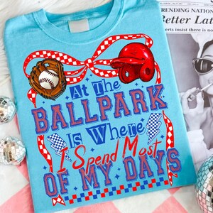Op de afbeelding: Een lichtblauw t-shirt met een afbeelding van een honkbalhandschoen, een honkbal en een rode slaghelm. De tekst "At The BALLPARK Is Where I Spend Most OF MY DAYS" staat in blauwe en rode letters, geaccentueerd door rood geblokte linten.