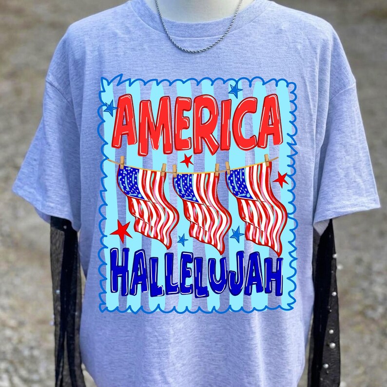 Puede incluir: Una camiseta gris claro con un estampado gr&aacute;fico que presenta el texto rojo AMERICA sobre tres ilustraciones de banderas estadounidenses colgadas en un tendedero, y el texto azul HALLELUJAH debajo, enmarcado por un borde festoneado azul claro.