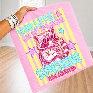 May include: Pink blanket with a raccoon wearing sunglasses and holding a drink. Text reads "YOUR LITTLE RAY OF SARCASTIC SUNSHINE HAS ARRIVED" in blue and yellow. The design includes stars and yellow stripes.
