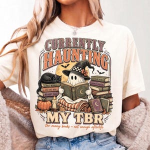 Pode incluir: Uma t-shirt cor de creme com um gr&aacute;fico de um fantasma com chap&eacute;u de bruxa, lendo um livro enquanto est&aacute; sentado numa manta. O design inclui pilhas de livros, um gato preto, uma ab&oacute;bora, uma vela e o texto "CURRENTLY HAUNTING MY TBR".