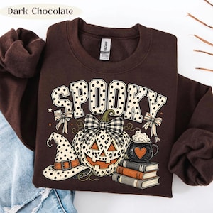May include: A dark chocolate brown crewneck sweatshirt featuring a graphic print with the word SPOOKY in white polka-dot letters. The design includes a leopard print pumpkin with a plaid bow, a polka-dot witch hat, a stack of books, and a mug.