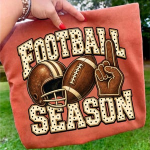 Puede incluir: Un bolso de tela color naranja quemado con un estampado gr&aacute;fico que incluye el texto FOOTBALL SEASON en una fuente color crema con puntos negros. El dise&ntilde;o muestra un casco de f&uacute;tbol americano, un bal&oacute;n y un dedo de espuma marr&oacute;n.