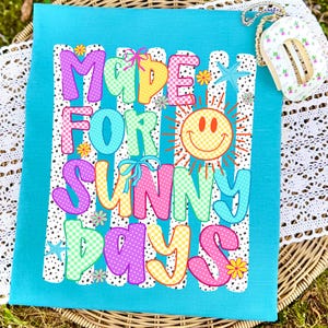 May include: A bright turquoise t-shirt featuring a graphic print with the text MADE FOR SUNNY DAYS in colorful, patterned bubble letters. The design includes a yellow smiley face sun, small blue starfish, and tiny flowers over white vertical stripes.