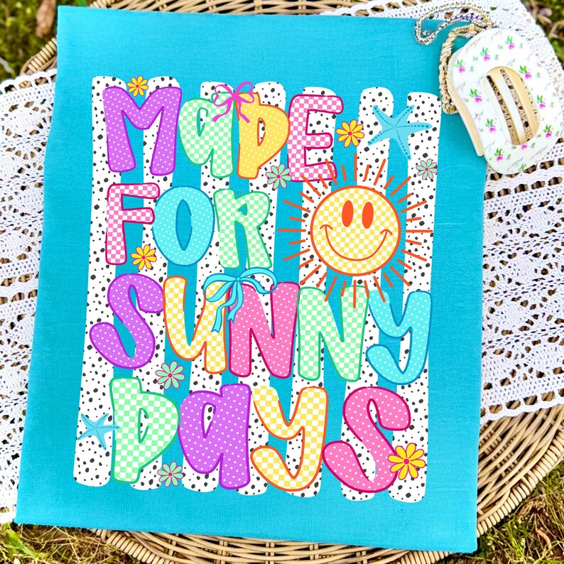 May include: A bright turquoise t-shirt featuring a graphic print with the text MADE FOR SUNNY DAYS in colorful, patterned bubble letters. The design includes a yellow smiley face sun, small blue starfish, and tiny flowers over white vertical stripes.