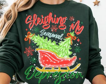 May include: Dark green sweatshirt with a festive graphic. The design features a red sleigh carrying two green Christmas trees, snowflakes, and the text "Sleighing My Seasonal Depression" in red and green script.