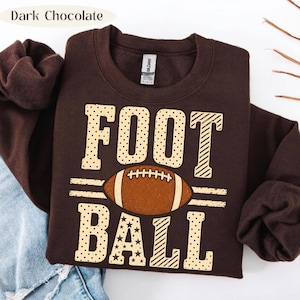 May include: A dark chocolate brown crewneck sweatshirt featuring the text FOOT BALL in large, light tan letters with various patterns including dots, stripes, and stars. A brown American football graphic is centered between the two words.