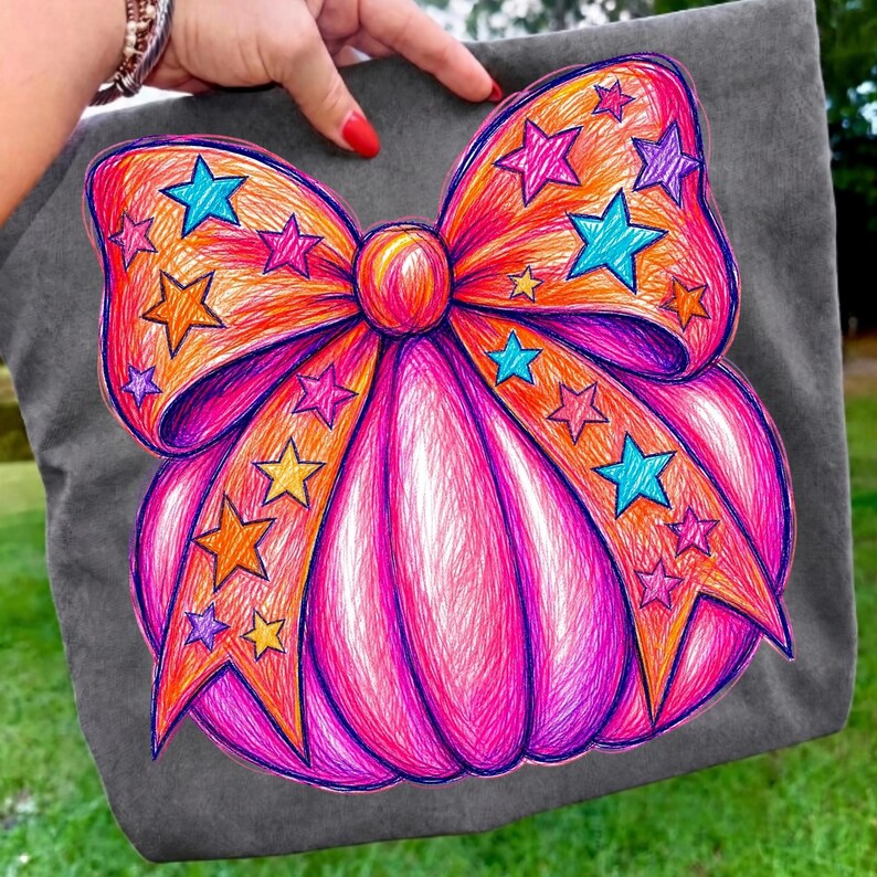 May include: A dark gray fabric tote bag featuring a large, vibrant graphic of a pink pumpkin adorned with a prominent orange bow. The design is filled with colorful scribbled lines and scattered star shapes in shades of blue, yellow, and purple.